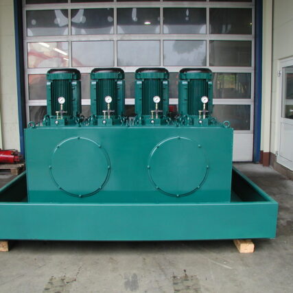 Großes grünes Hydraulikaggregat mit vier senkrecht montierten Elektromotoren und Druckanzeigen, platziert auf einer Auffangwanne in einer Werkstatthalle.