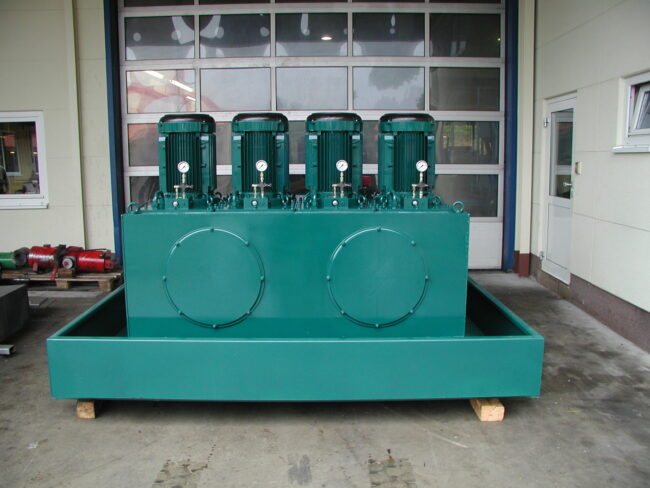 Großes grünes Hydraulikaggregat mit vier senkrecht montierten Elektromotoren und Druckanzeigen, platziert auf einer Auffangwanne in einer Werkstatthalle.