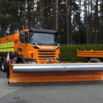 Ein orangefarbener Scania-LKW mit großem Schneepflug an der Front steht bereit für den Winterdiensteinsatz. Auf der Ladefläche befindet sich ein Streuaufsatz.