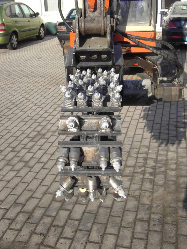 Frontalaufnahme eines hydraulischen Grabenfräskopfes an einem Bagger. Zu sehen sind zahlreiche Meißelzähne auf mehreren Walzen, die zum Fräsen von Boden und Asphalt eingesetzt werden.