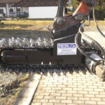 Seitenansicht eines am Baggerarm montierten Grabenfräskopfes mit umlaufender Kette und Meißelwerkzeugen. Die hydraulischen Leitungen und der Antriebsmotor sind deutlich sichtbar.