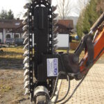 Der Grabenfräskopf steht in vertikaler Arbeitsstellung. Die umlaufende Meißelkette, der hydraulische Motor und die Verbindung zum Baggerarm sind klar erkennbar.