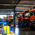 Innenaufnahme einer Werkstatt, in der mehrere orangefarbene Winterdienst-LKW gewartet oder umgebaut werden. Mitarbeitende arbeiten an hochgekippten Fahrzeugkabinen.