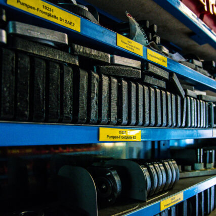 Mehrere Reihen dunkler Pumpen-Frontplatten und Metallteile liegen ordentlich sortiert in einem Regal mit gelben Beschriftungsschildern.