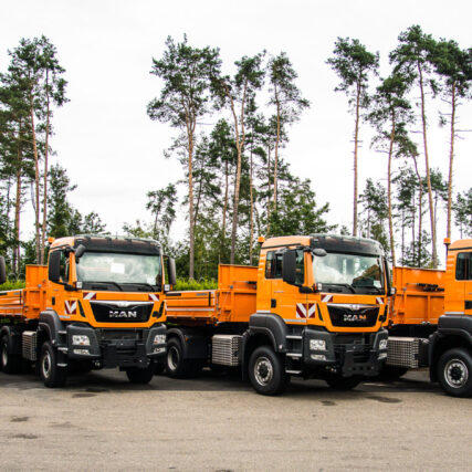 Reihe orangefarbener MAN-LKW für Winterdienst und kommunale Einsätze, geparkt vor einer Baumreihe. Die Fahrzeuge sind einheitlich für Straßenunterhalt ausgestattet.