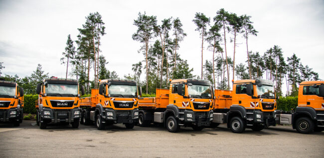 Reihe orangefarbener MAN-LKW für Winterdienst und kommunale Einsätze, geparkt vor einer Baumreihe. Die Fahrzeuge sind einheitlich für Straßenunterhalt ausgestattet.
