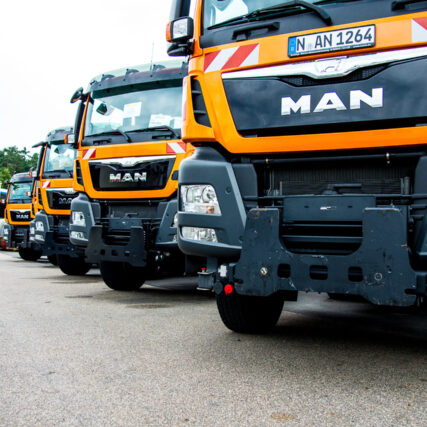 Ein MAN-LKW mit großen Schneepflugmodulen an der Front steht auf einem Betriebshof. Fahrzeug und Pflug sind in typischer Winterdienst-Signallackierung gestaltet.